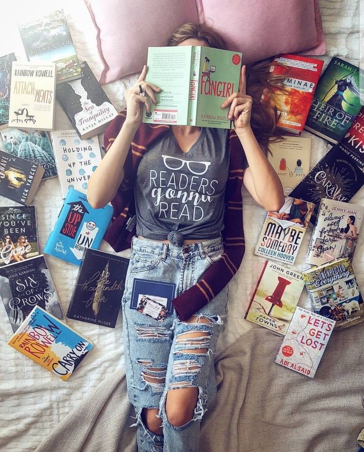 a woman laying in bed reading a book with lots of books on the floor behind her