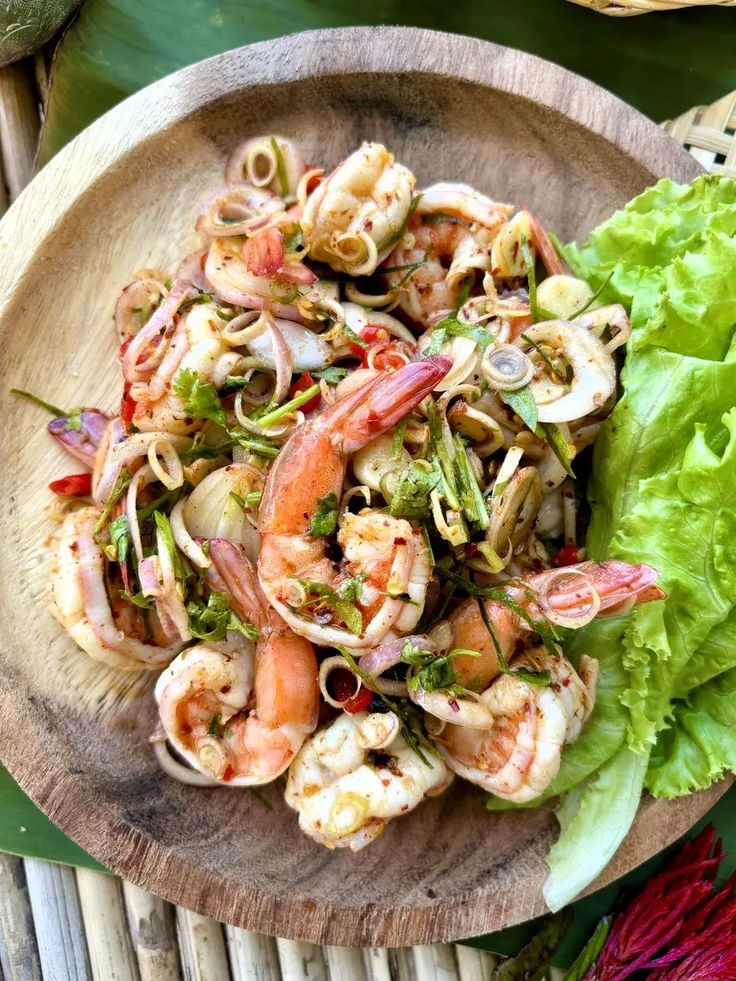 a plate filled with shrimp and veggies on top of a wooden table next to a green leafy salad