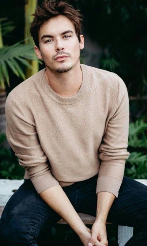 a man sitting on top of a wooden bench wearing a brown sweater and black jeans