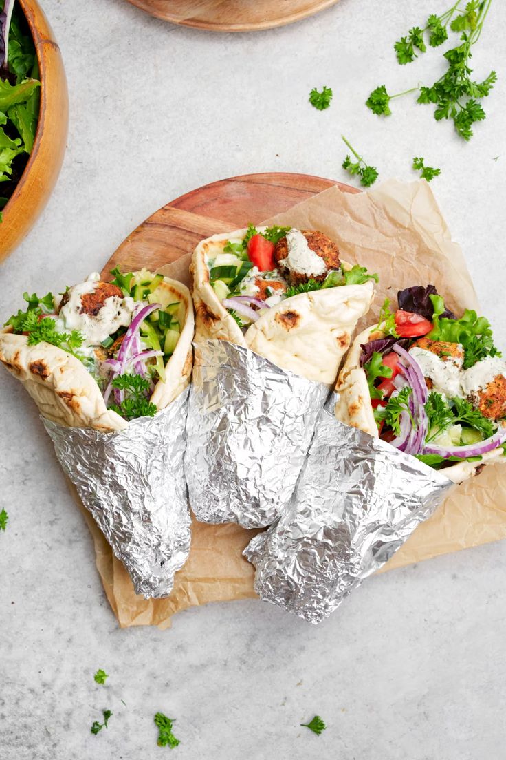 three burritos wrapped in aluminum foil on a cutting board with salad and lettuce