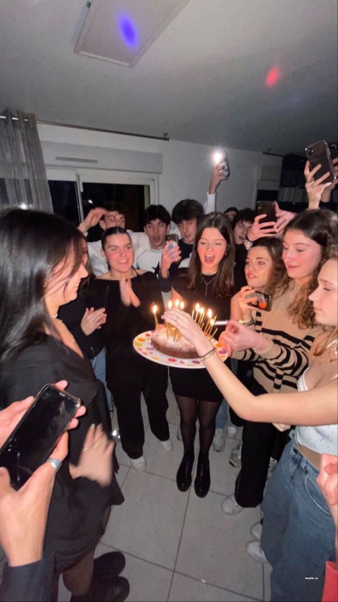 a group of people standing around a cake with lit candles on it and one person holding a cell phone up to the camera