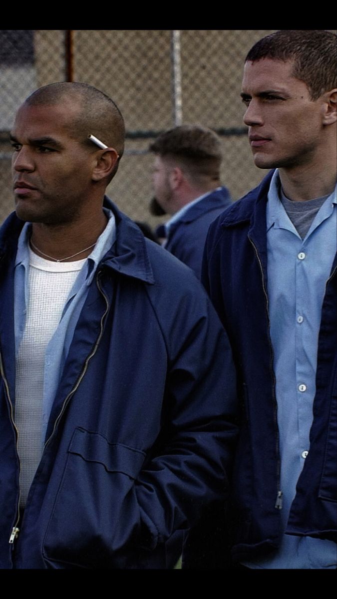 two men standing next to each other in front of a chain link fence, one wearing a blue jacket