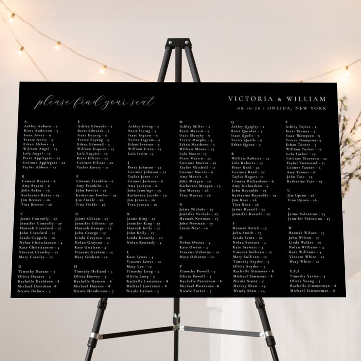 a black and white wedding seating chart on a easel with lights in the background