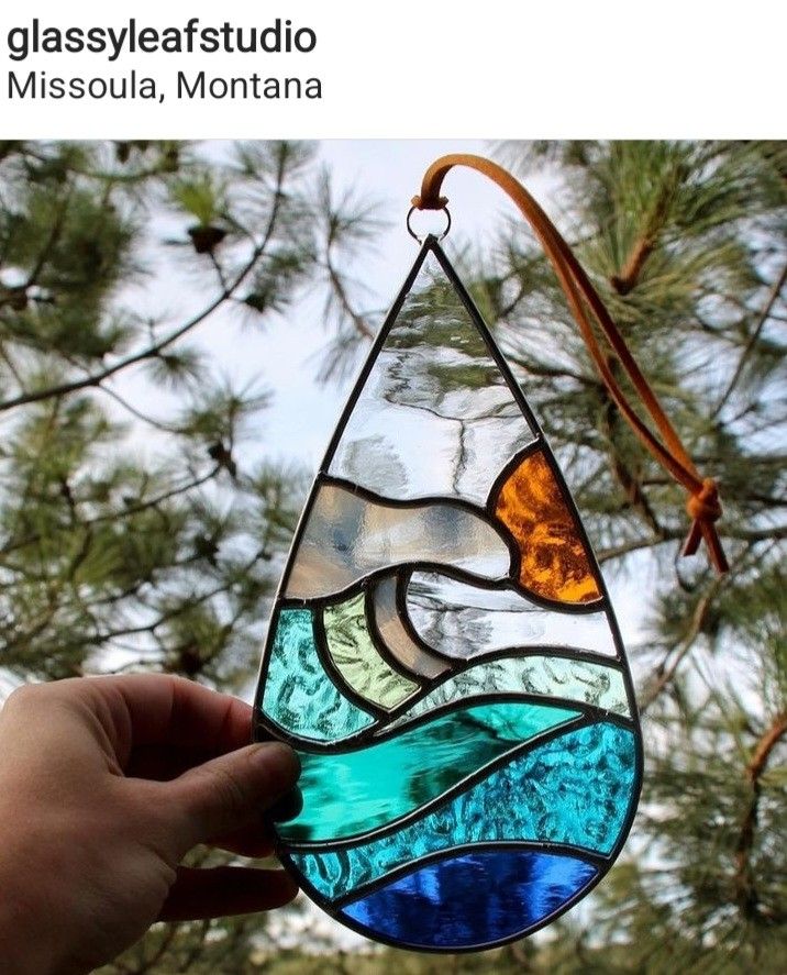 a hand holding up a stained glass hanging from a tree with water and waves in it