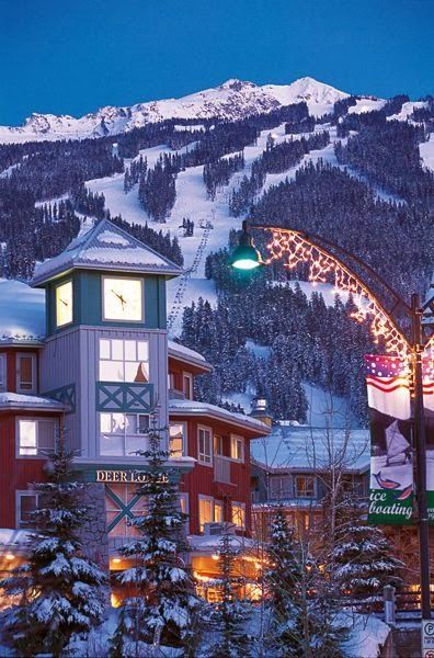 the town is lit up at night with christmas lights on it's buildings and snow covered mountains in the background