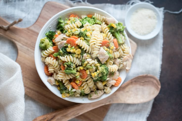 a white bowl filled with pasta salad next to a wooden spoon on top of a cutting board