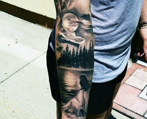 a man with a tattoo on his arm is standing in front of the water and trees