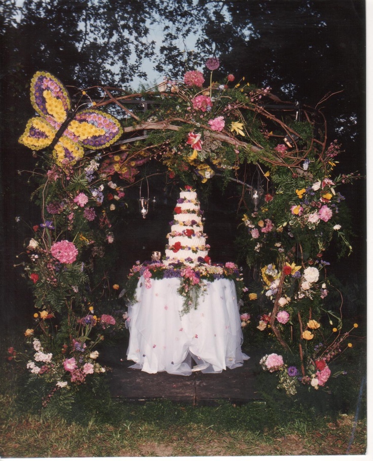 a wedding cake with flowers and butterflies on it in the middle of a flower arch