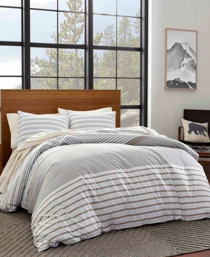 a bed with white and grey striped comforter in front of large windows next to a wooden headboard