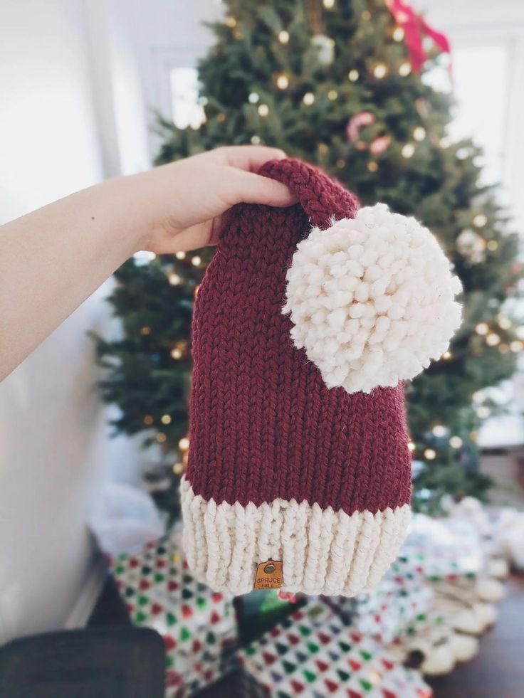 a hand holding a red and white knitted mitt next to a christmas tree