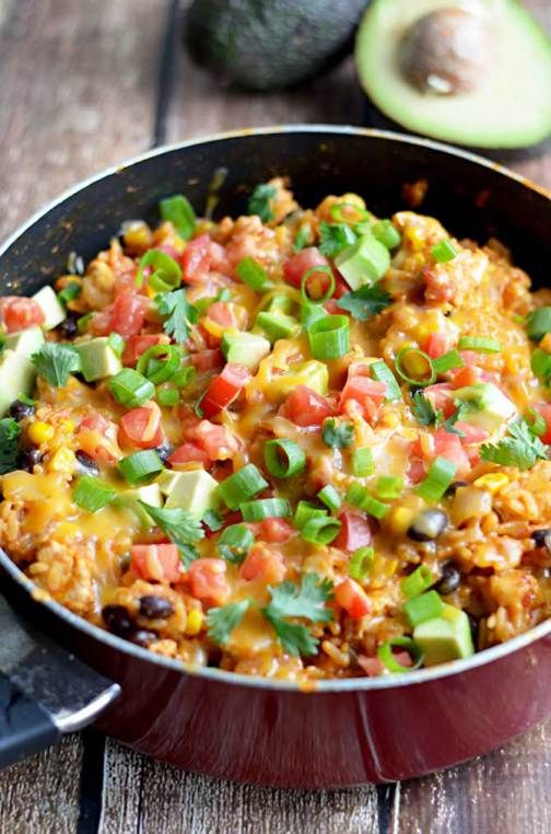 a skillet filled with rice and vegetables on top of a wooden table next to an avocado