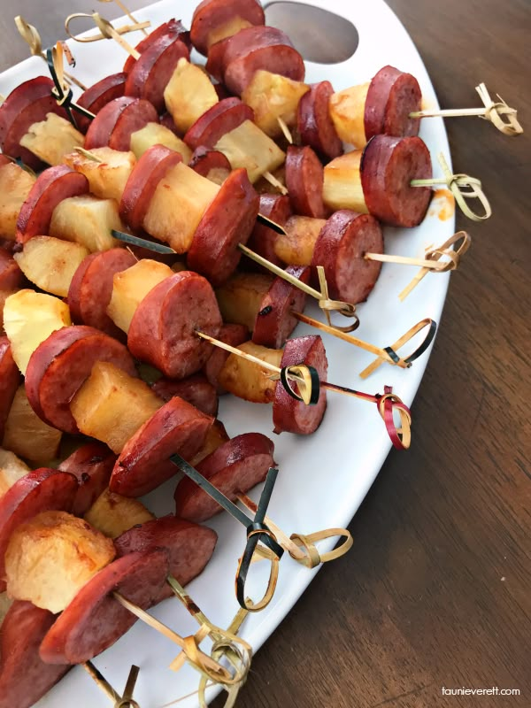 several skewered sausages and pineapple on a white platter with toothpicks