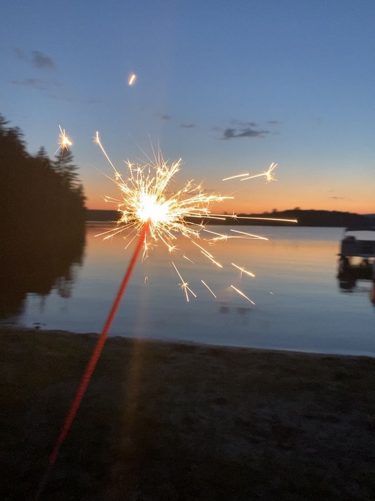 a sparkler is lit up in the air near water and trees at sunset or dawn