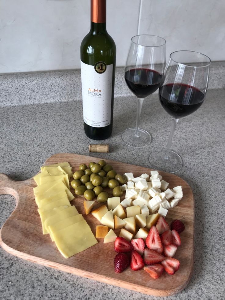 a wooden cutting board topped with cheese and fruit next to two glasses of red wine