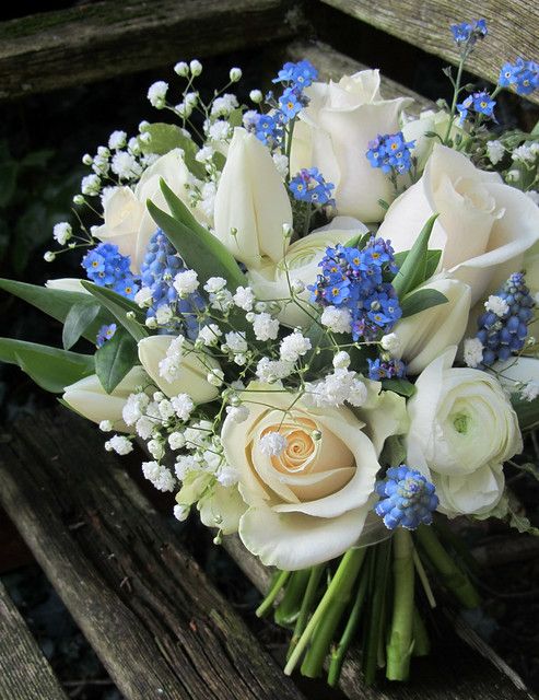 a bouquet of white roses and blue flowers on a wooden bench with greenery in the background