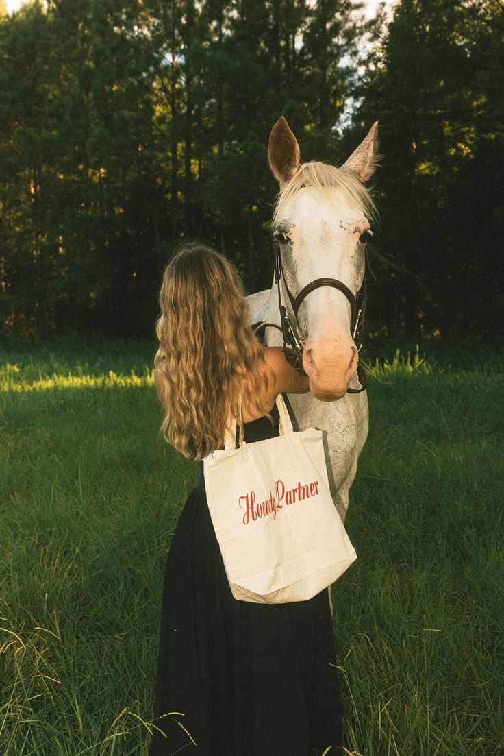 a woman is standing in the grass next to a horse with a shopping bag on it's back