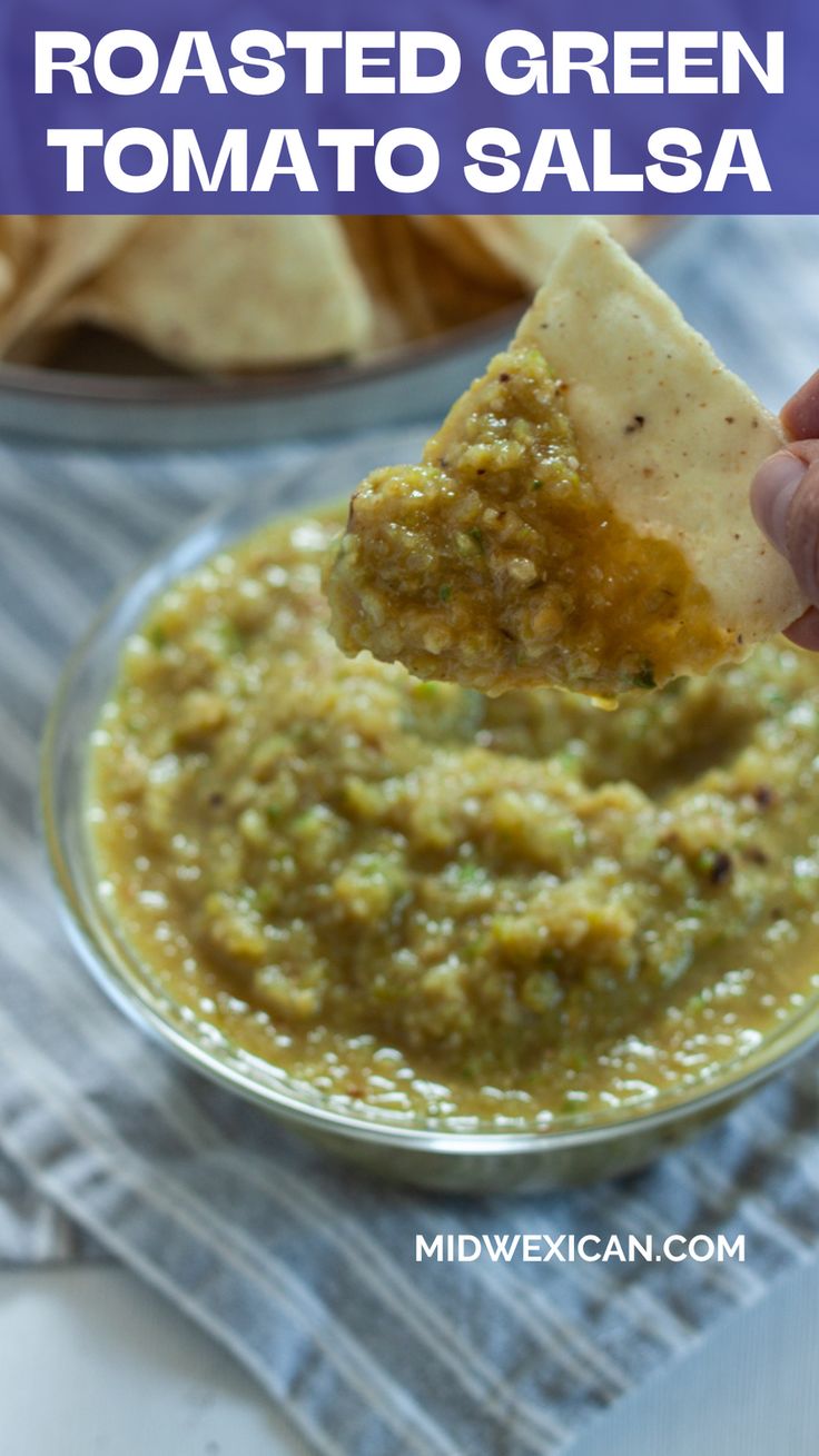 a hand holding a tortilla chip over a bowl of roasted green tomato salsa