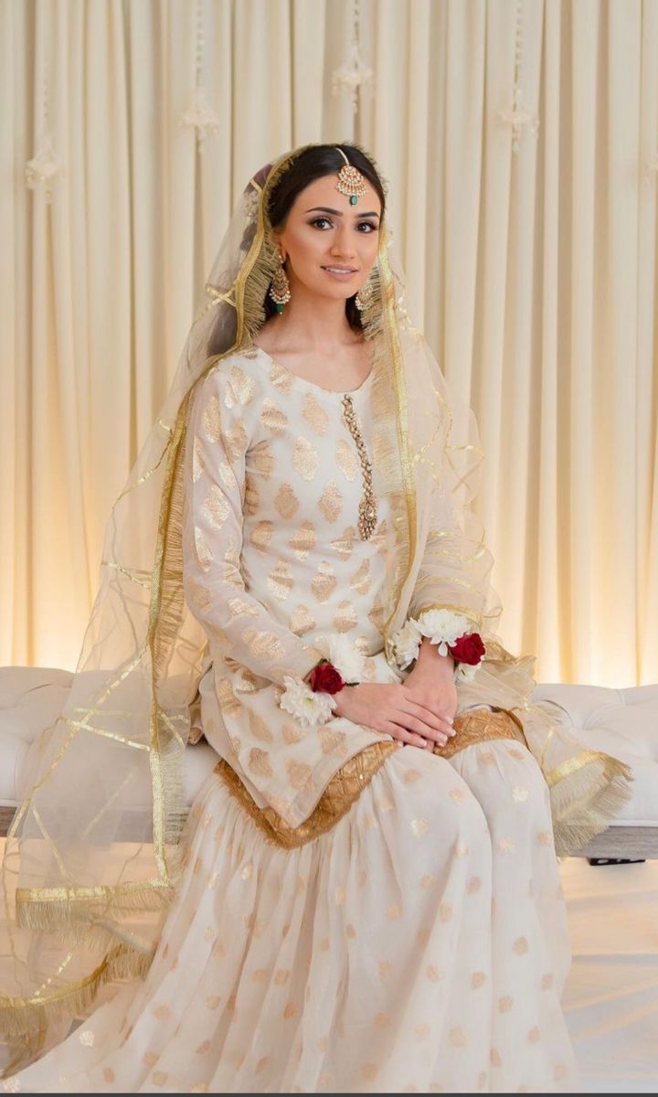 a woman sitting on top of a bed wearing a white dress with gold trims