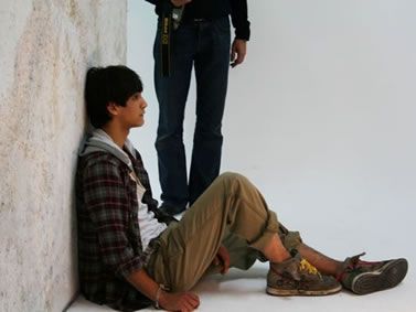 two young men sitting next to each other in front of a white wall and one man standing