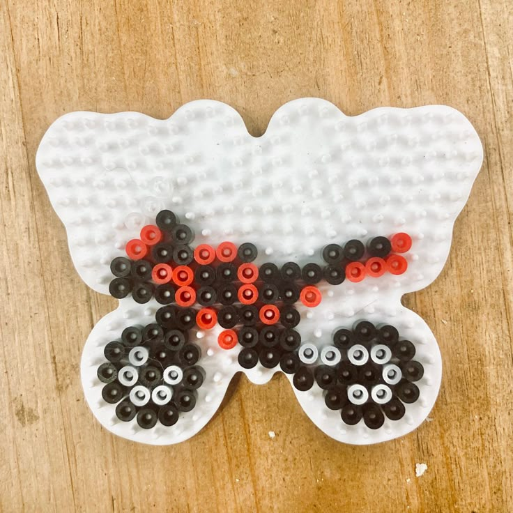 a white and black butterfly made out of plastic beads on a wooden surface with red eyes