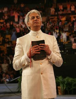 an older man in a white suit holding a book and looking at the camera while standing on stage