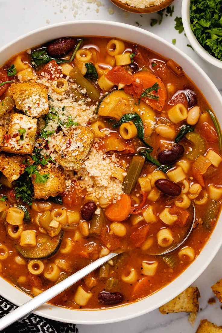 a white bowl filled with pasta and vegetable soup on top of a table next to bread