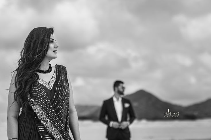 a man and woman standing next to each other near the ocean with mountains in the background