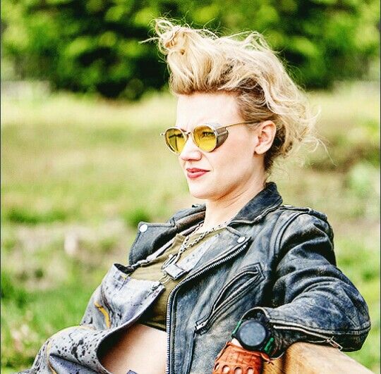 a woman wearing sunglasses sitting on top of a wooden bench next to a field and trees