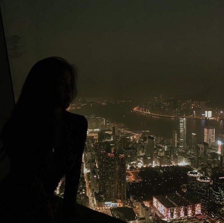 a woman sitting on top of a tall building looking at the city lights in the distance