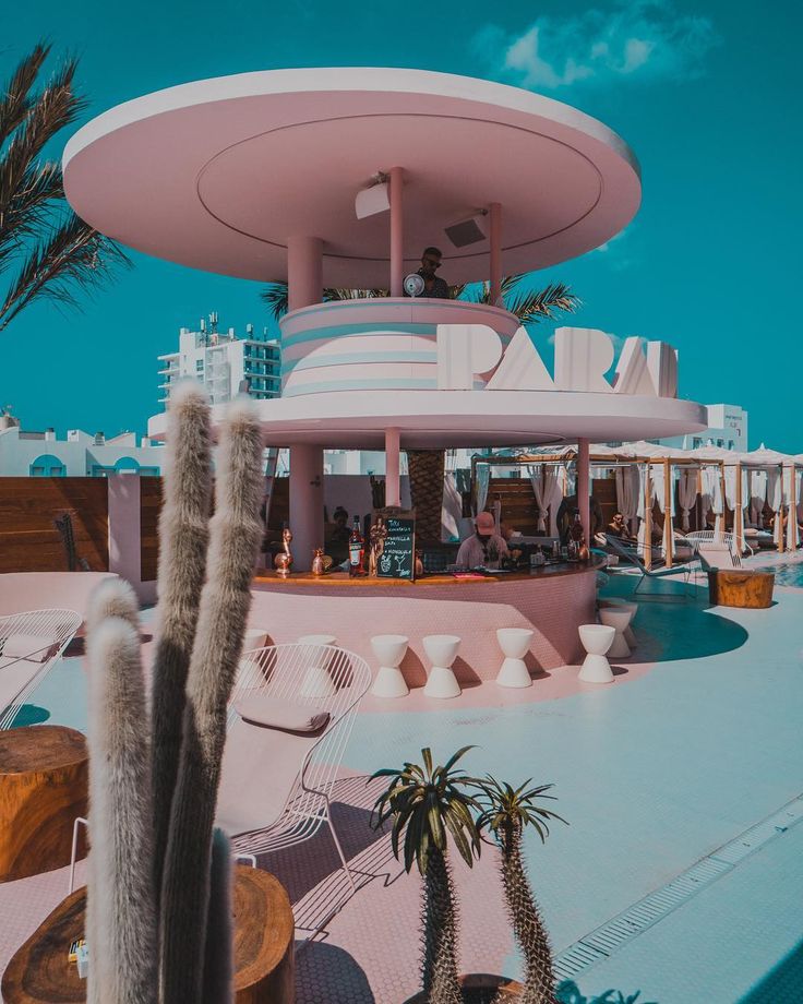 a pink building with palm trees in the foreground and people sitting at tables on the other side