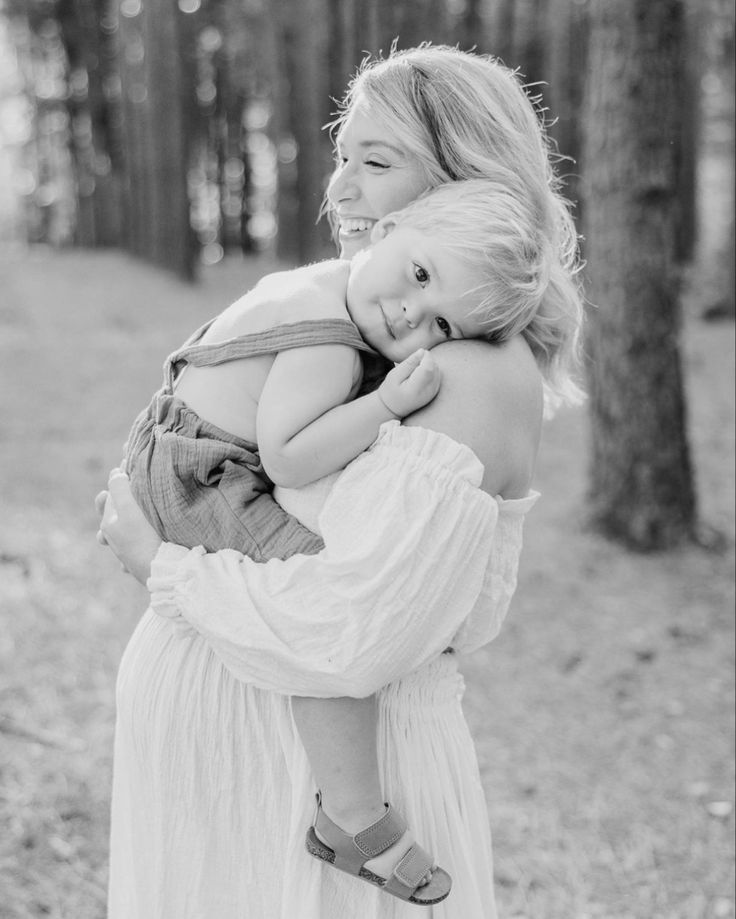 a woman holding a baby in her arms and smiling at the camera with trees behind her