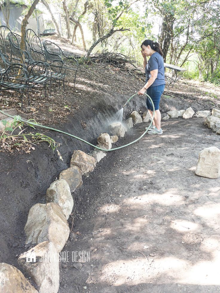 a woman is using a hose to clean the soil in her garden area with rocks