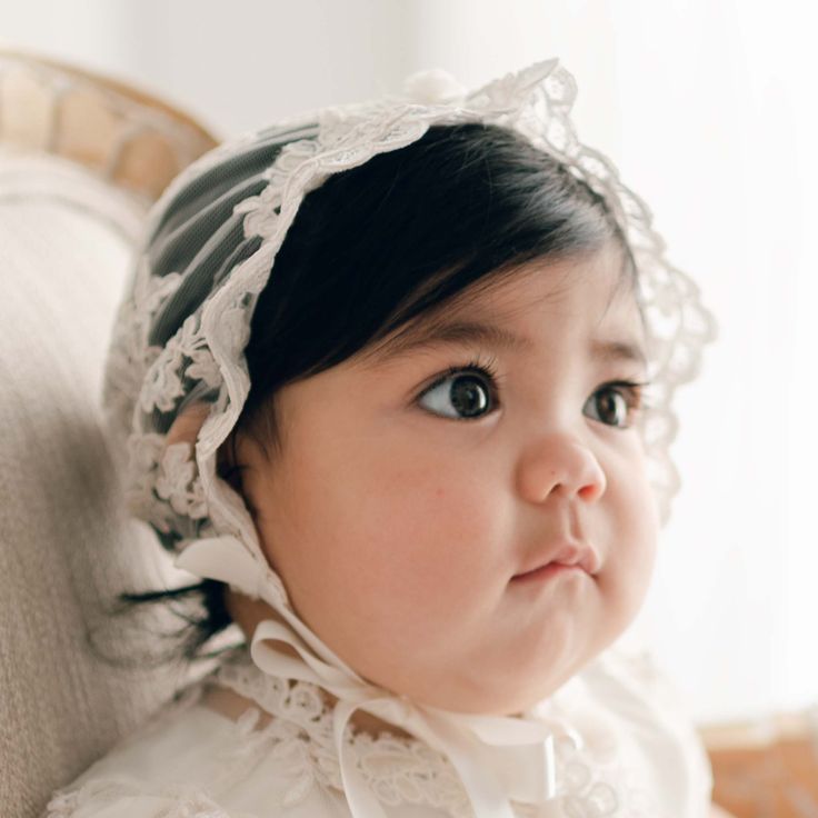 Baby girl with dark hair wearing the Penelope Lace Bonnet and matching baptism dress gazes away to the side of the camera. Elegant Organza Lace For Ceremony, Elegant Lace Veil For Ceremony, White Adjustable Lace Bonnet, White Lace Adjustable Bonnet, Fitted White Bonnet With Lace Trim, Elegant Lace Bonnet With Lace Trim, Elegant Adjustable Cream Bonnet, Lace Veil With Lace Trim For Ceremonies, Elegant Tulle Lace With Lace Trim