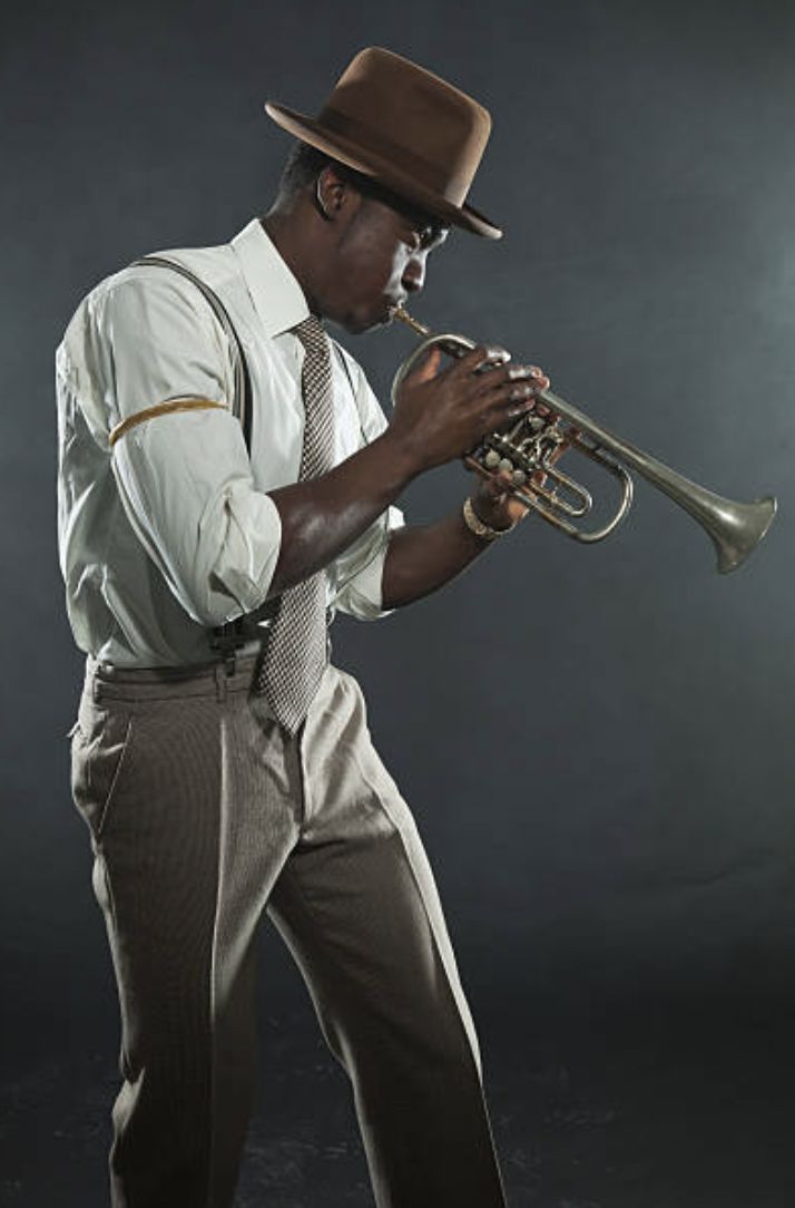 a man wearing a hat and holding a trumpet in his right hand while standing against a dark background