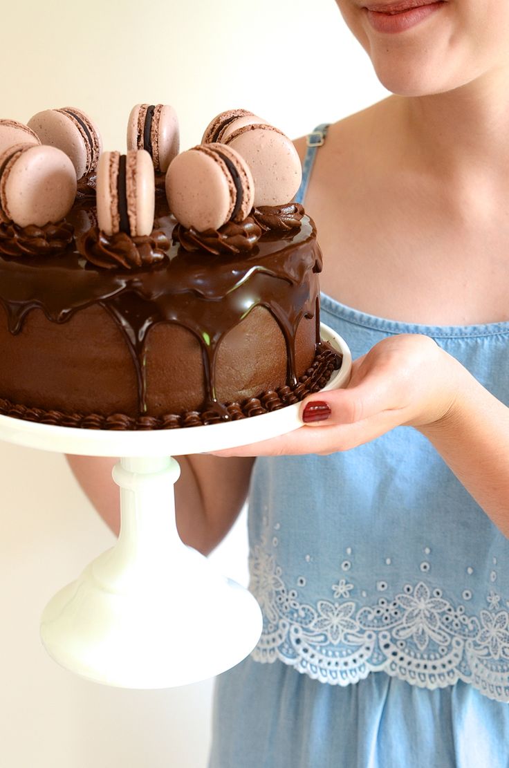 a woman holding a cake with chocolate frosting and macaroons on top in front of her