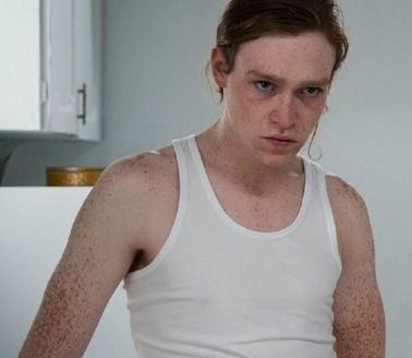 a young man in a tank top is looking at the camera while standing in front of a refrigerator