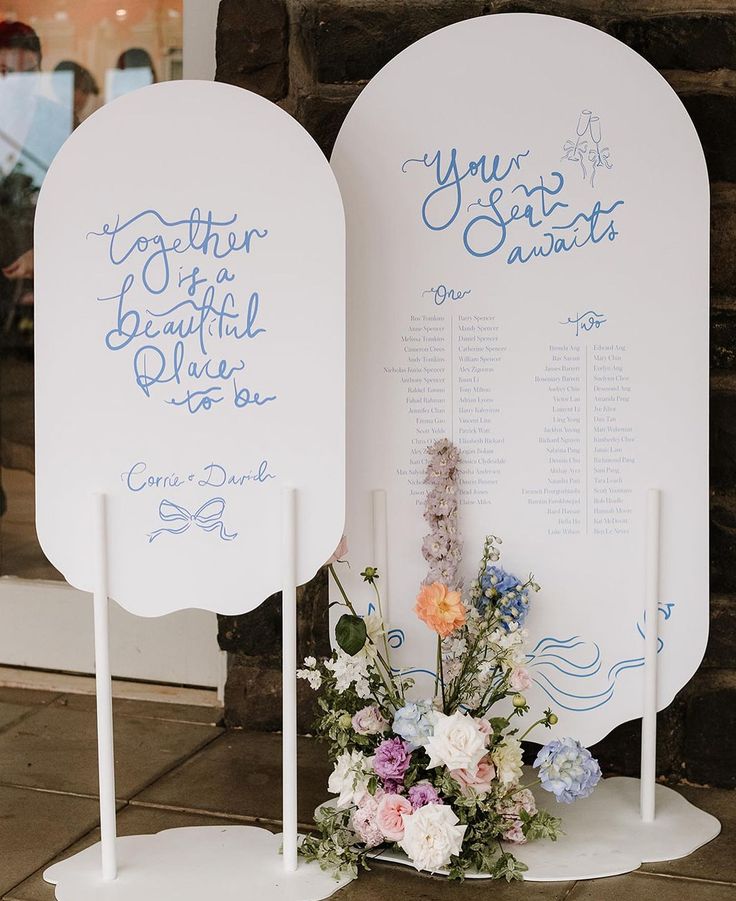 two white signs sitting next to each other with blue writing on them and flowers in the foreground