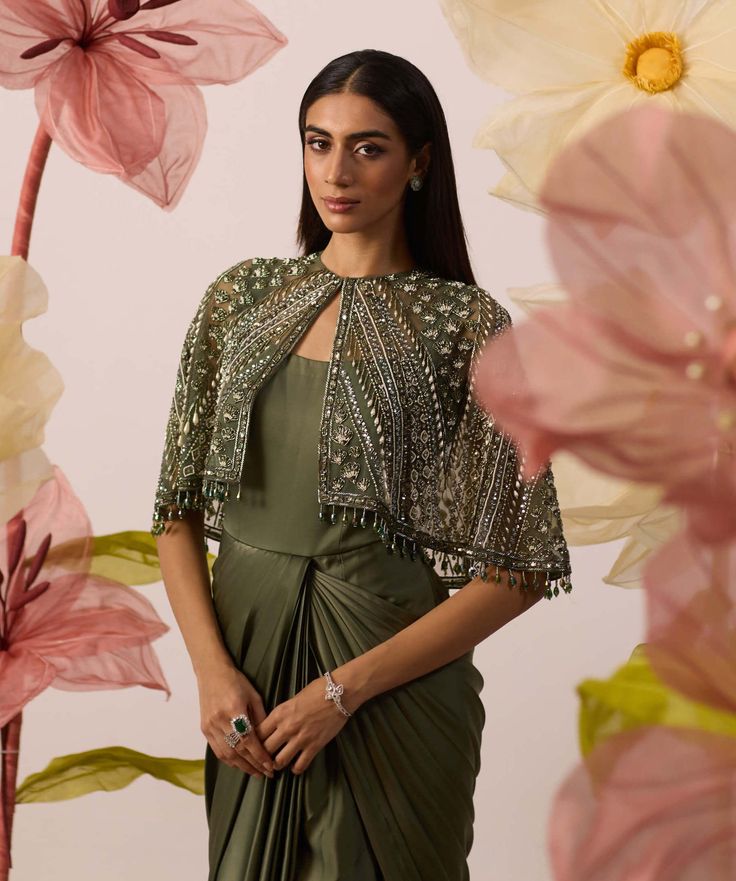 Display absolute finesse in this olive green drape set crafted in modal satin fabric. Featuring a long attached drape set and embroidered cape jacket with pearls & sequins, cutdana & mirror work, it's a picture-perfect wardrobe addition! Layer it up with perfect diamond jewelry and you're good to dazzle at the next occasion. Evening Draped Gown With Zari Work, Silk Dress With Draped Sleeves For Designer Wear, Silk Designer Dress With Draped Sleeves, Designer Silk Dress With Draped Sleeves, Anarkali Gown With Cape Sleeves For Evening, Festive Gown With Traditional Draped Sleeves, Festive Gown With Draped Sleeves, Fitted Gown With Dupatta And Cape Sleeves, Green Cape Sleeves Sets For Eid