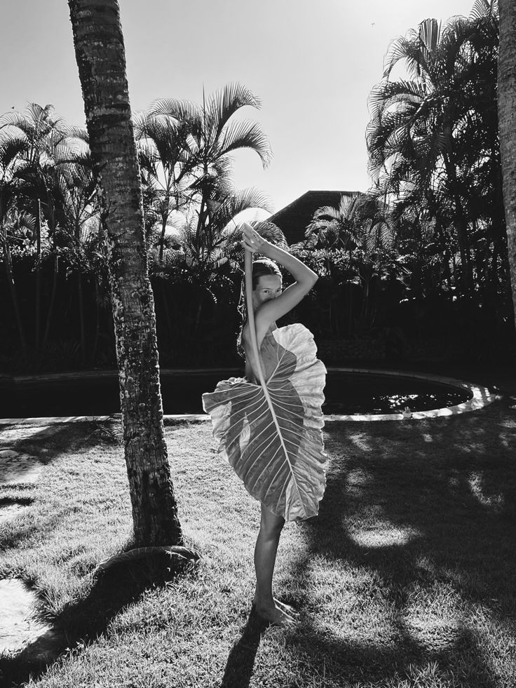 a woman standing in the grass near two palm trees with her arms behind her head