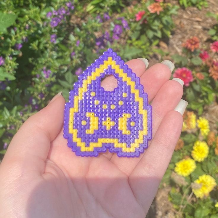 a hand holding a purple and yellow beaded brooch in front of some flowers