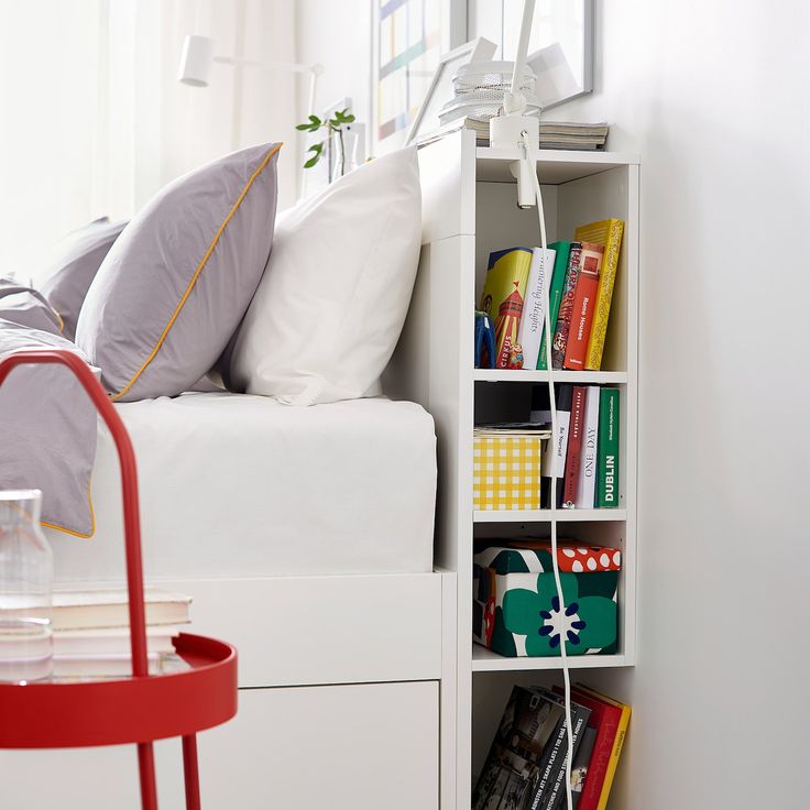 a white bed sitting next to a red table with books on it's shelves