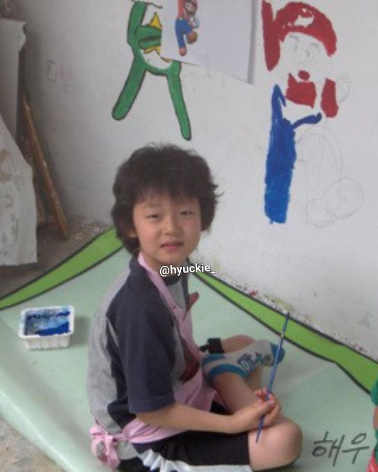 a little boy sitting on the ground with some paintbrushes in his hand and another child's drawing behind him