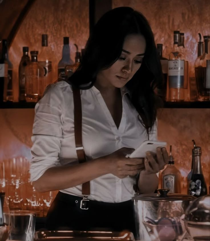 a woman standing in front of a bar with bottles on the counter and looking at her cell phone