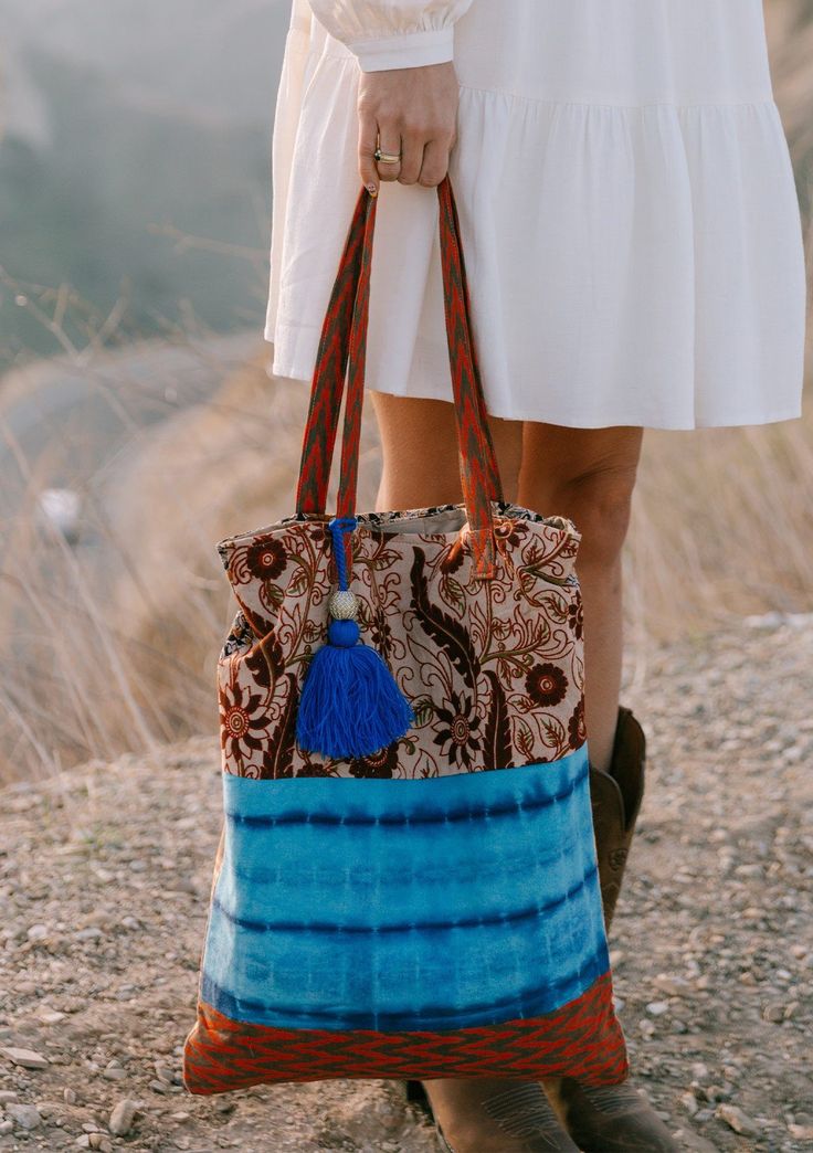 [Color: Natural/Blue] A woman standing outside on a cliff wearing a white dress and holding a bohemian tie dye patchwork tote bag. Featuring a large tassel accent. Patchwork Hobo Tote Bag, Bohemian Multicolor Canvas Bag, Bohemian Cotton Shoulder Bag For Market, Bohemian Fabric Shoulder Bag For Daily Use, Brown Bohemian Fabric Bags, Bohemian Brown Fabric Bags, Bohemian Multicolor Canvas Bag For Everyday Use, Multicolor Bohemian Canvas Bag For Everyday, Bohemian Cotton Canvas Shoulder Bag