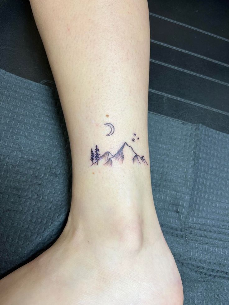 a small tattoo on the ankle of a woman with mountains and trees in the background