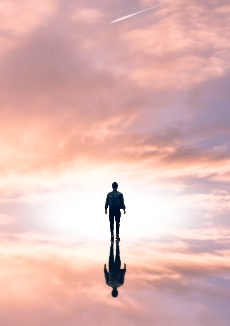 a man standing in the middle of a body of water under a pink sky with an airplane flying overhead