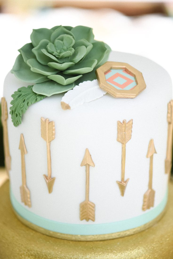 a white and gold cake with an arrow design on it, topped with a succulent plant