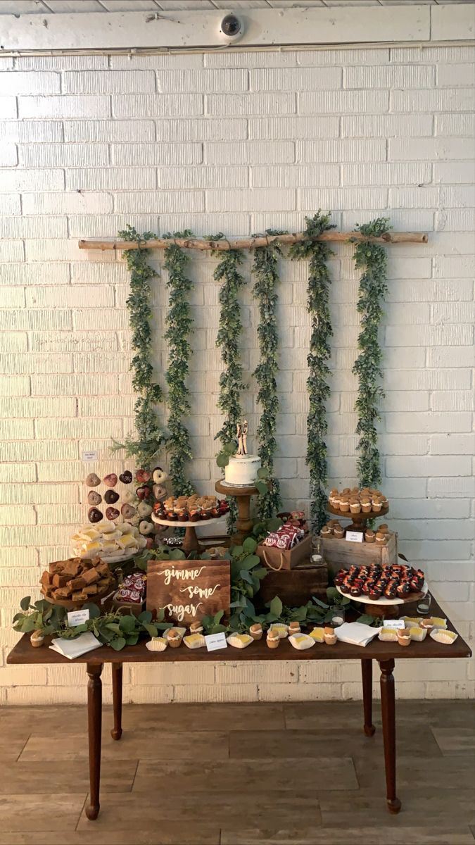 a table topped with lots of desserts next to a wall covered in greenery