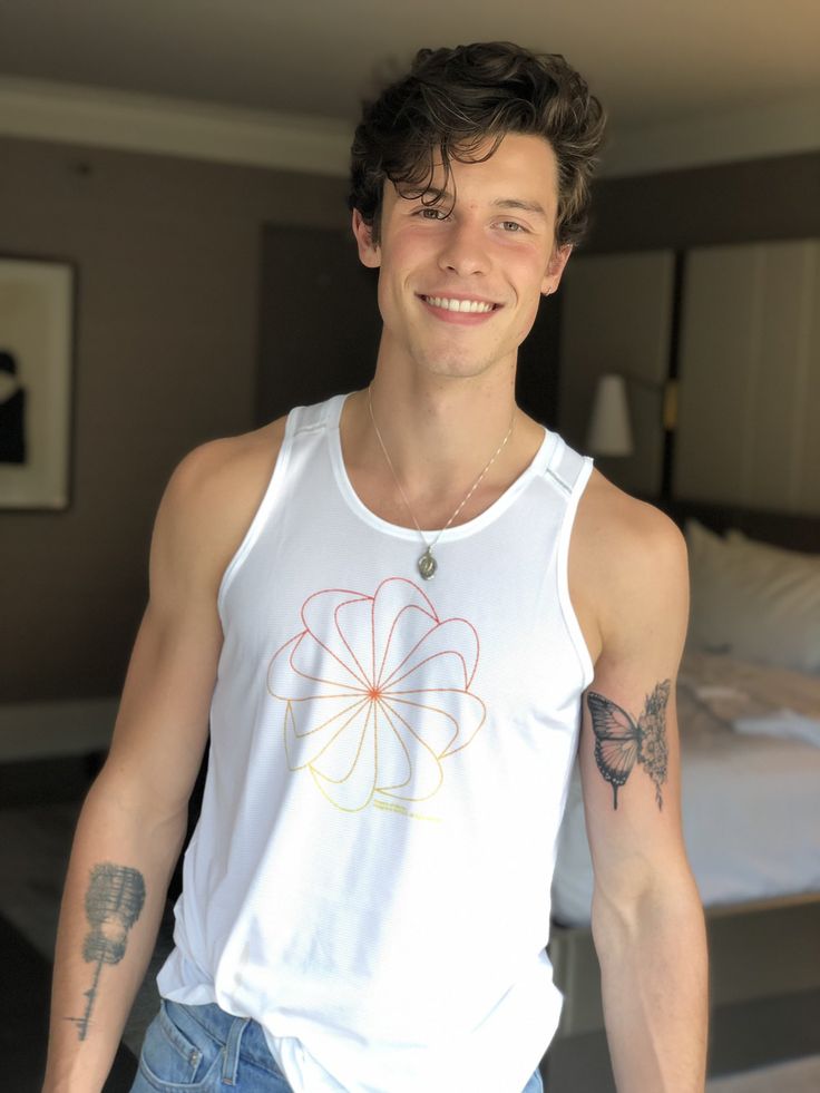 a man with tattoos standing in front of a bed wearing a white tank top and blue jeans