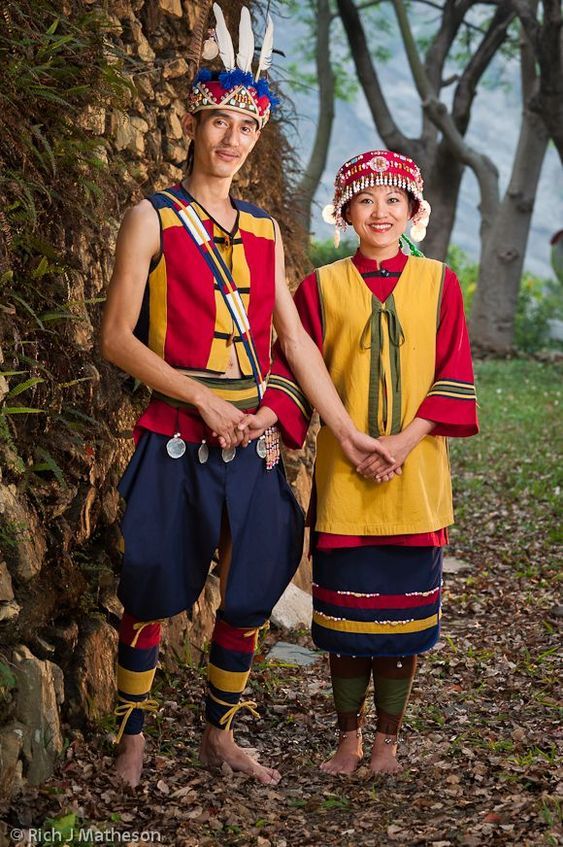 two people standing next to each other in colorful clothing and headdress, with trees behind them